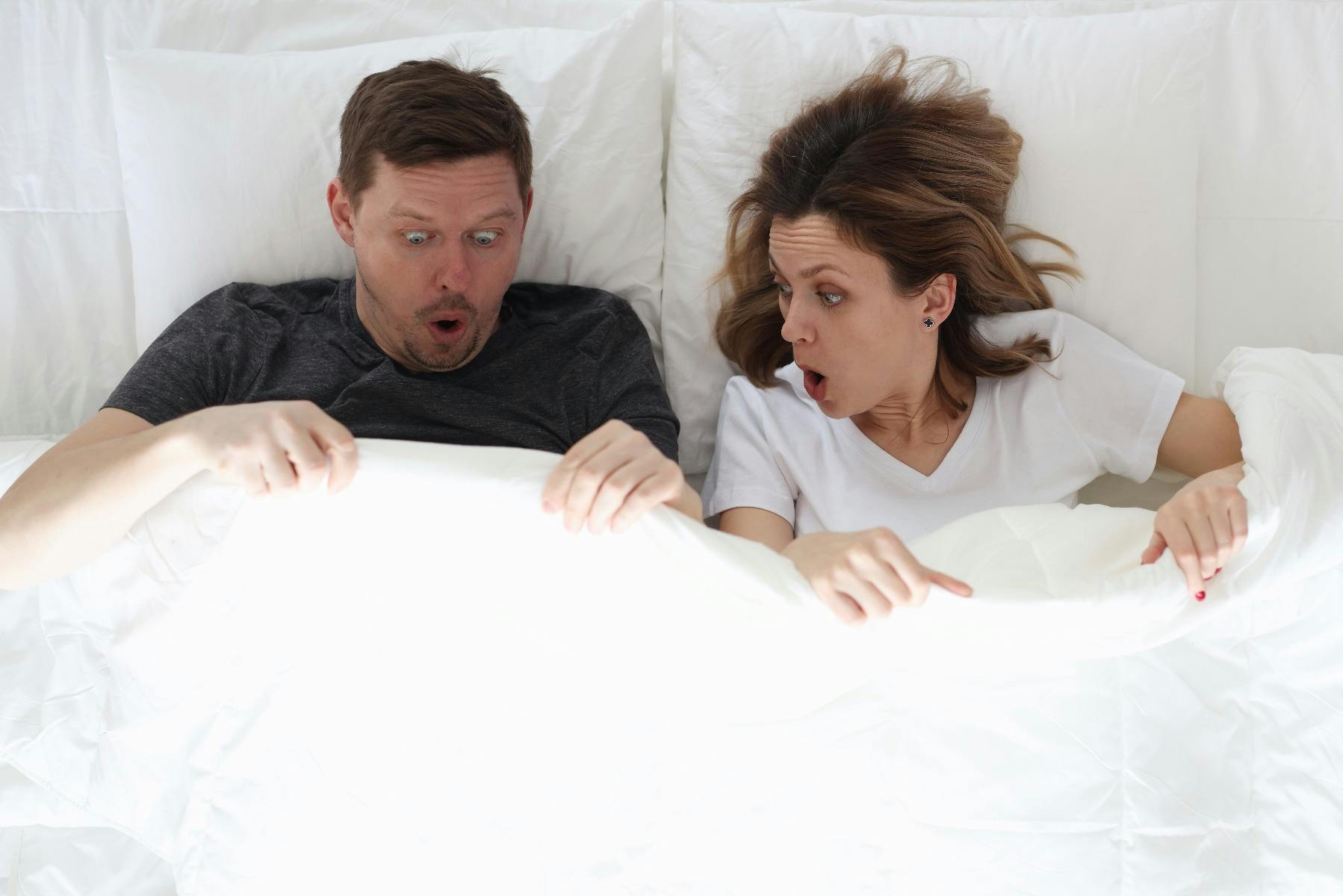 A heterosexual couple in bed with surprised expressions on there faces due to the man having a hard erection after taking Viagra for his erectile dysfunction