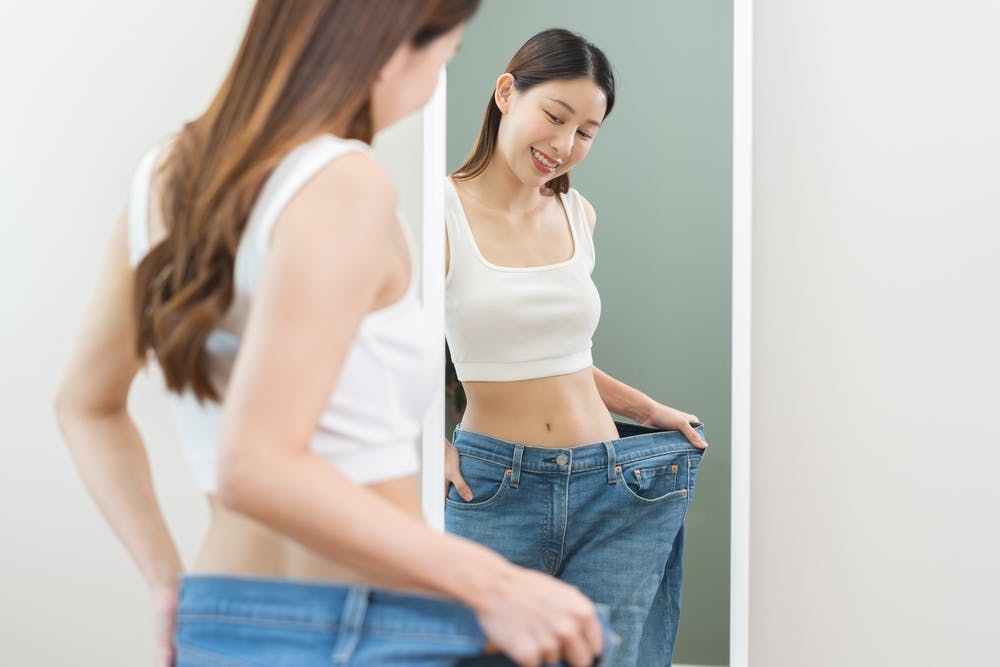 A lady looking into the mirror whilst holding a pair of loose jeans up because of how much weight she has lost